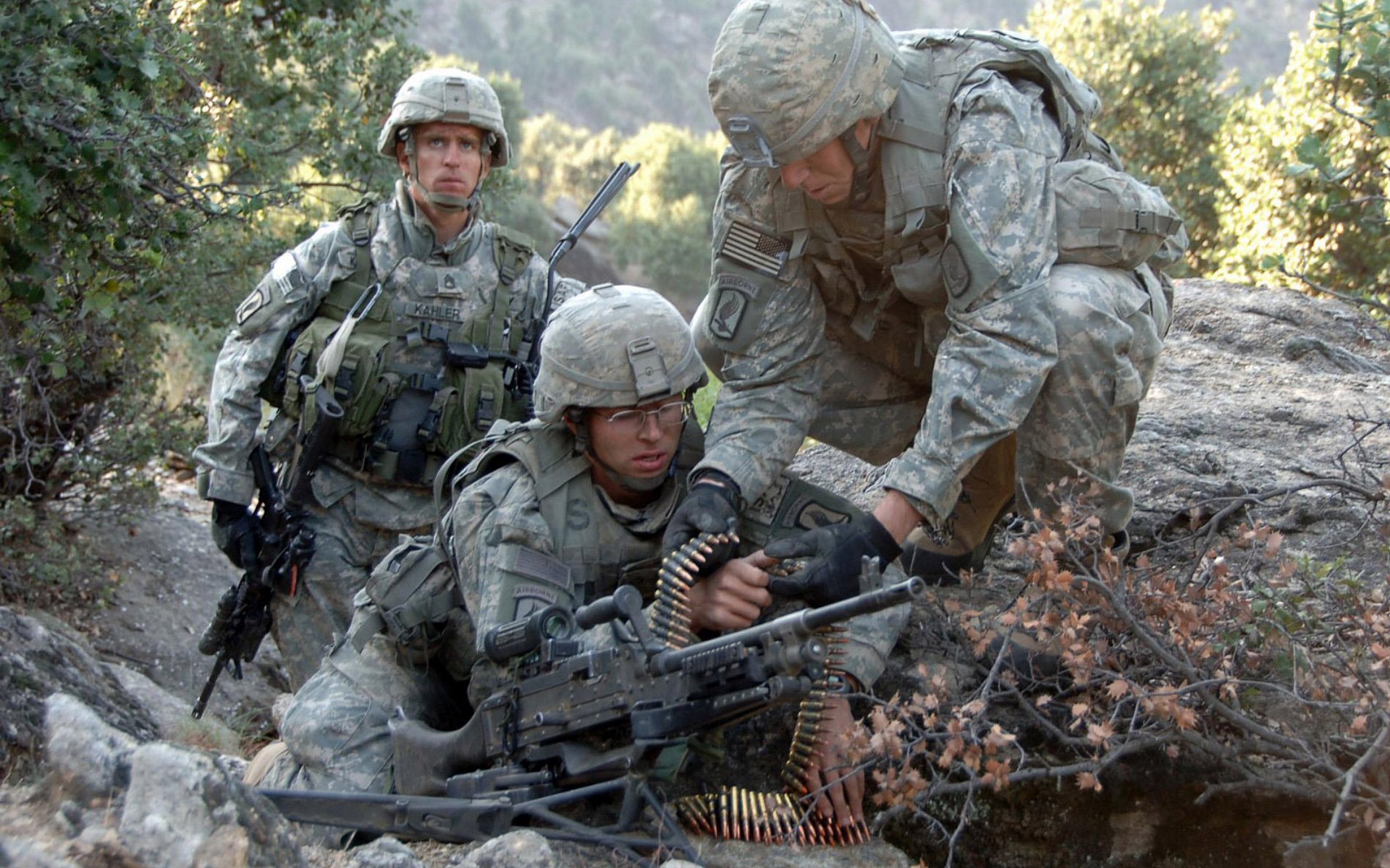 HD desktop wallpaper featuring three soldiers in full military gear operating a machine gun in a forested outdoor setting.