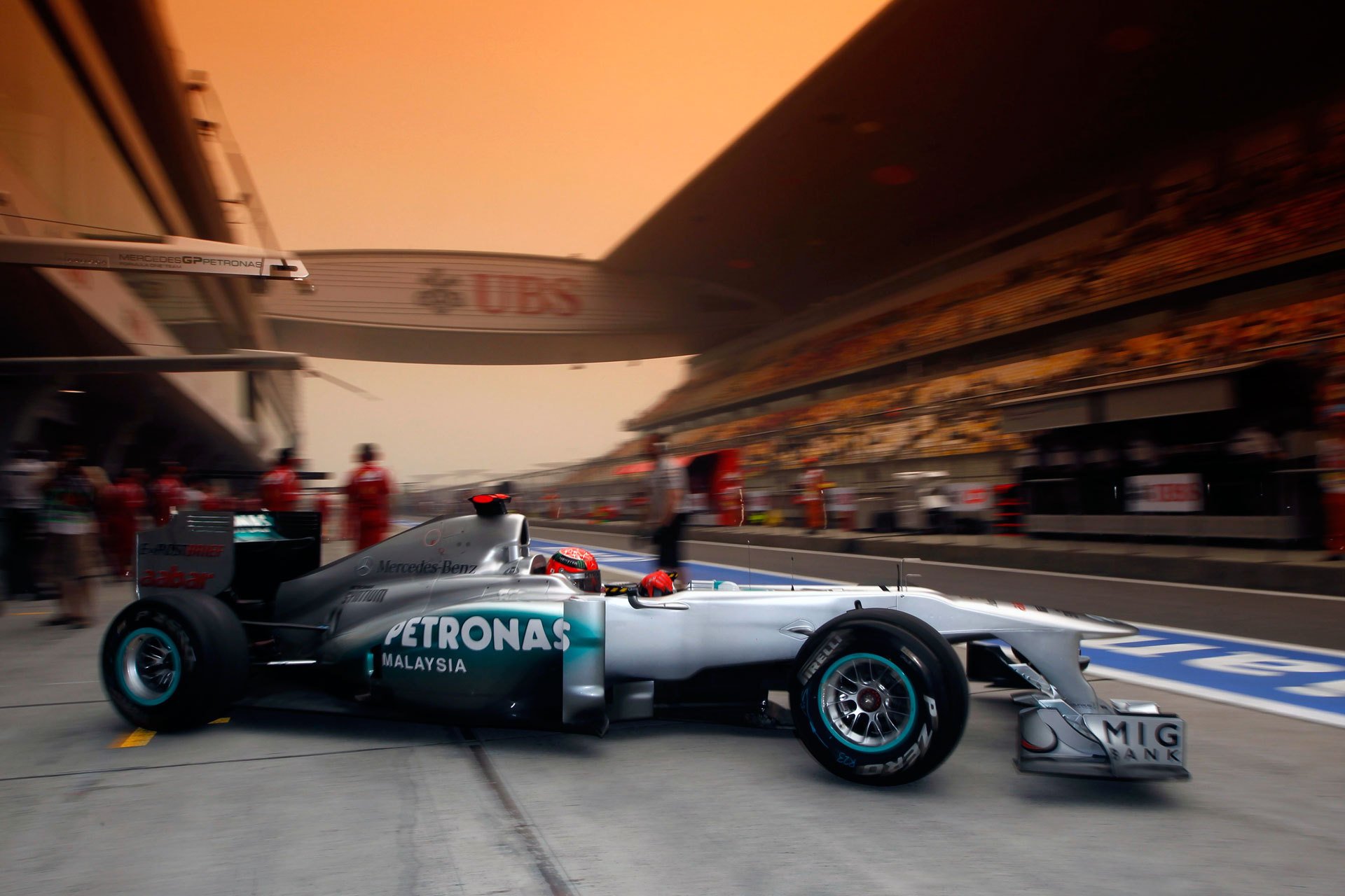HD PC desktop wallpaper background — F1 sports: Petronas Mercedes Formula 1 car in the pit lane, crew and grandstands under warm sunset light.
