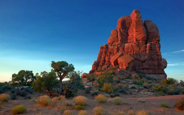 HD PC desktop wallpaper showcasing a striking red rock formation bathed in warm sunlight, surrounded by desert vegetation under a clear blue sky.