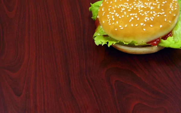 A close-up of a delicious hamburger with sesame seeds, lettuce, and ketchup, set against a rich wooden background, creating an appetizing HD desktop wallpaper.
