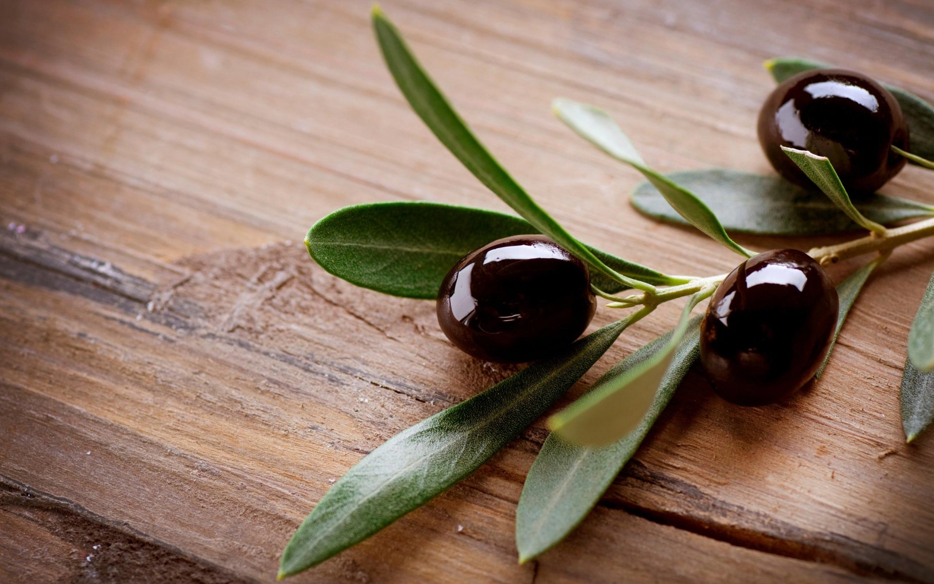 A close-up of three black olives with green leaves, resting on a rustic wooden surface, showcasing rich colors and natural textures. Suitable as a desktop wallpaper or background.