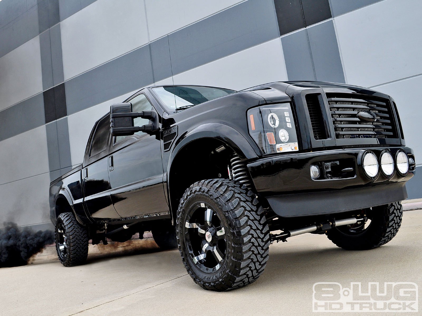 A sleek black truck with bold tires stands prominently against a modern building, showcasing its rugged design in this high-definition desktop wallpaper.