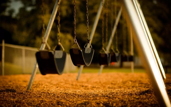 A close-up of empty swings lined up in a playground, set against a backdrop of soft sunlight filtering through trees, creating a serene atmosphere. HD photography for desktop wallpaper.