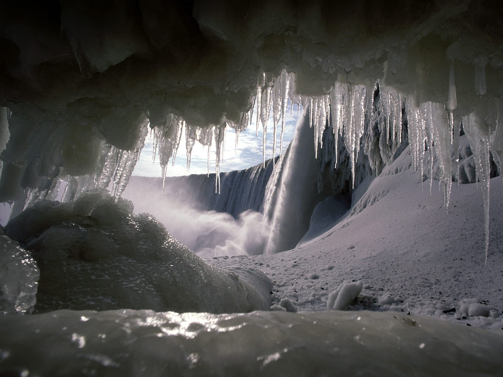 HD desktop wallpaper of a winter waterfall seen from inside a frozen cave. Snow and icicles frame the waterfall, blending elements of nature seamlessly.