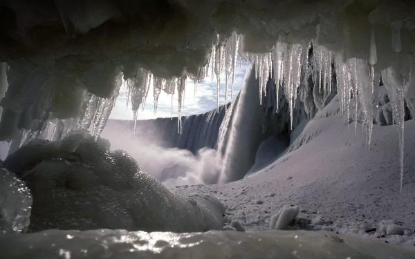 HD desktop wallpaper of a winter waterfall seen from inside a frozen cave. Snow and icicles frame the waterfall, blending elements of nature seamlessly.