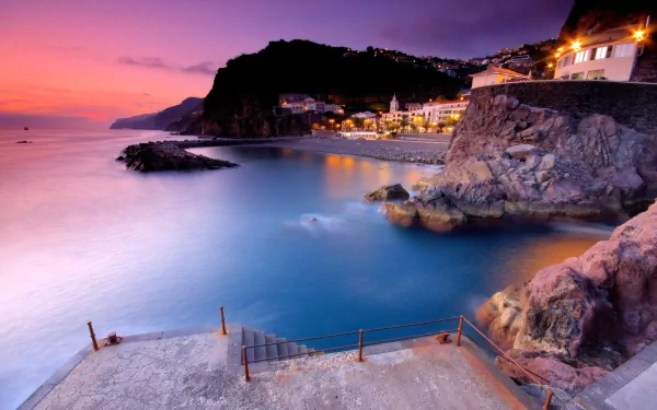 HD PC desktop wallpaper of a serene seascape at dusk, featuring calm waters, rocky cliffs, and illuminated coastal buildings under a vibrant purple and pink sky.