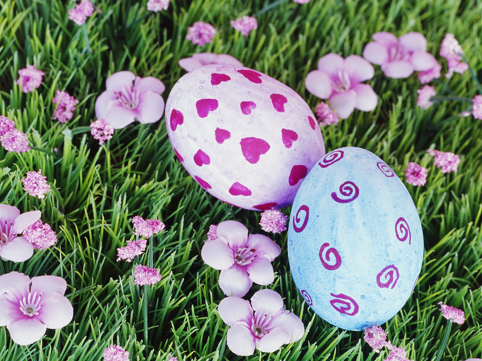 HD PC desktop wallpaper featuring two pastel Easter eggs with heart and swirl patterns nestled in green grass among pink flowers, capturing the holiday spirit.