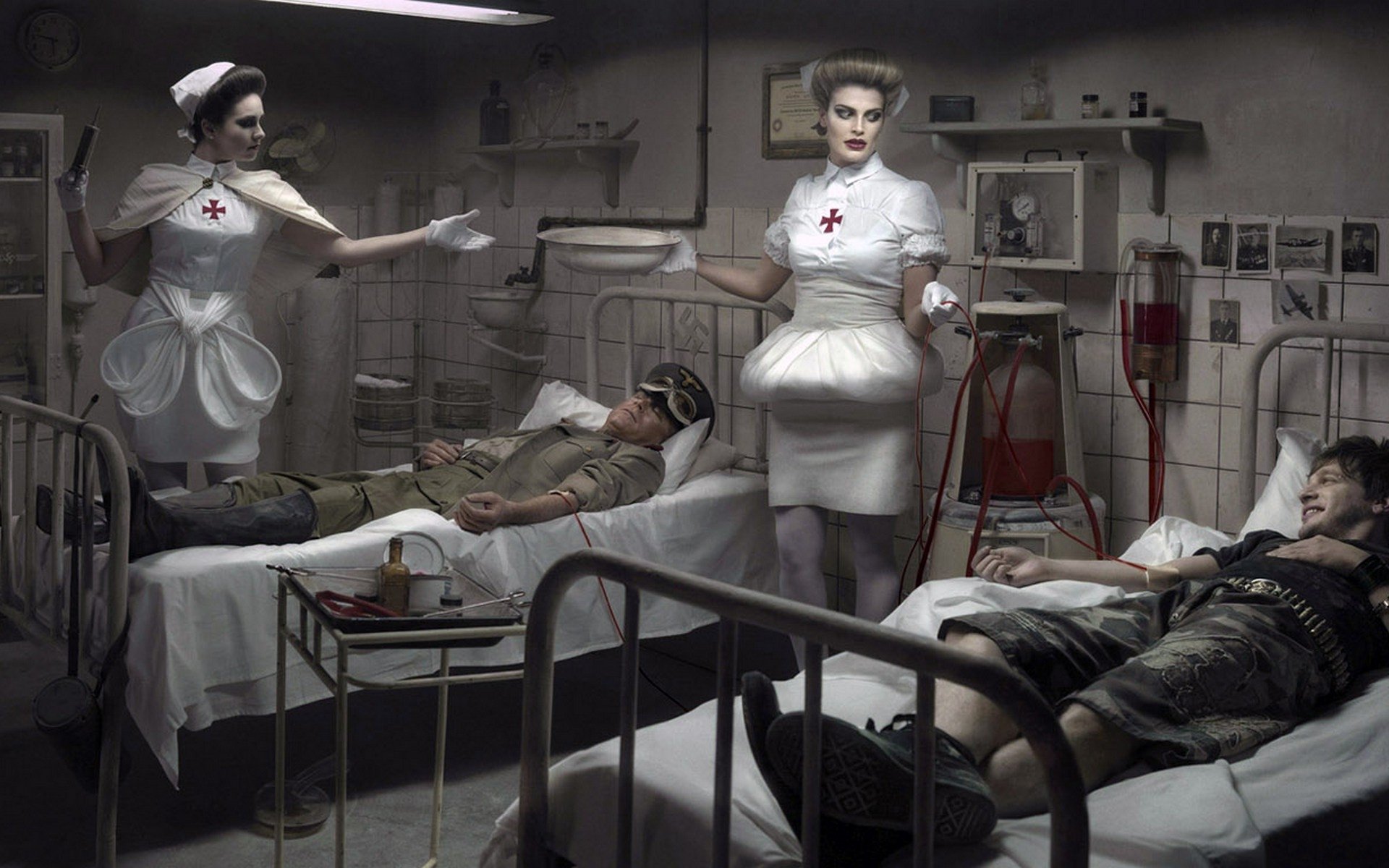 Dark HD PC desktop wallpaper background: a moody wartime hospital ward with two nurses in vintage uniforms tending wounded soldiers on metal beds under dim lights.