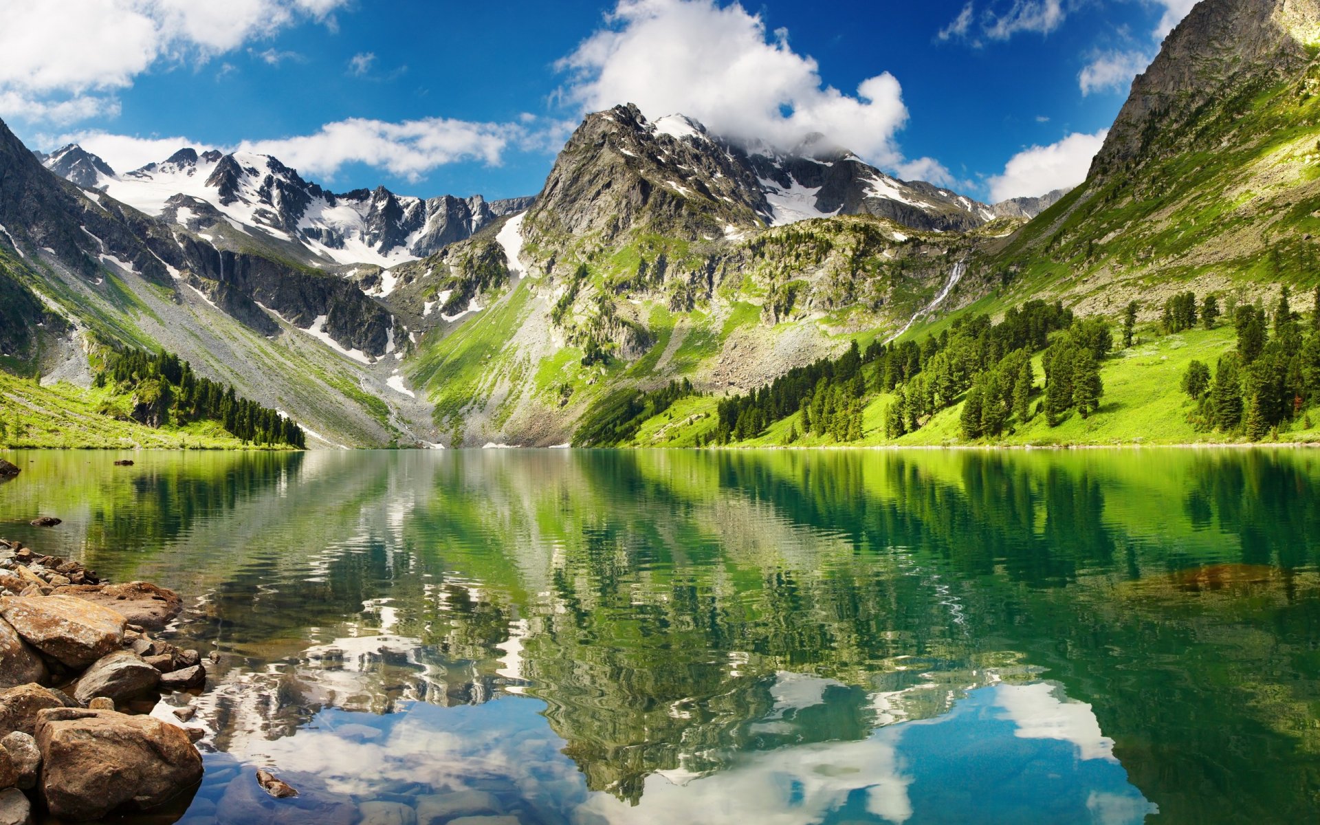 HD PC desktop wallpaper showcasing a vibrant mountain landscape reflected clearly in a calm lake, surrounded by lush greenery under a bright blue sky with scattered clouds.