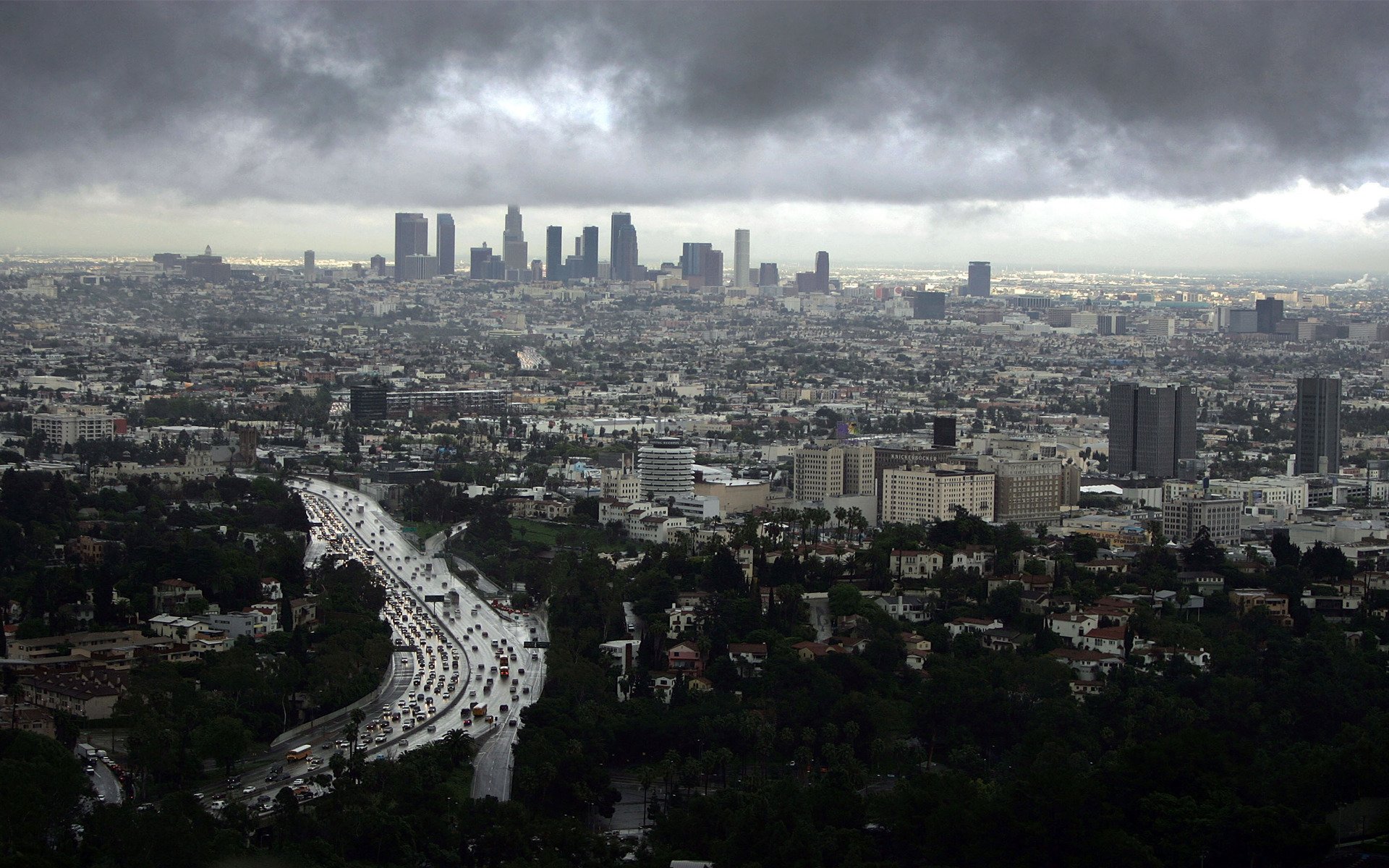 HD PC desktop wallpaper: man-made Los Angeles skyline and curving freeway seen from above beneath heavy storm clouds, wide urban panorama.