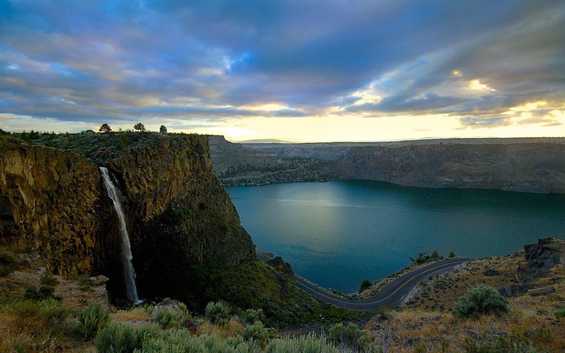 HD desktop wallpaper showcasing a stunning landscape photograph of a waterfall cascading into a serene lake under a dramatic cloudy sky.