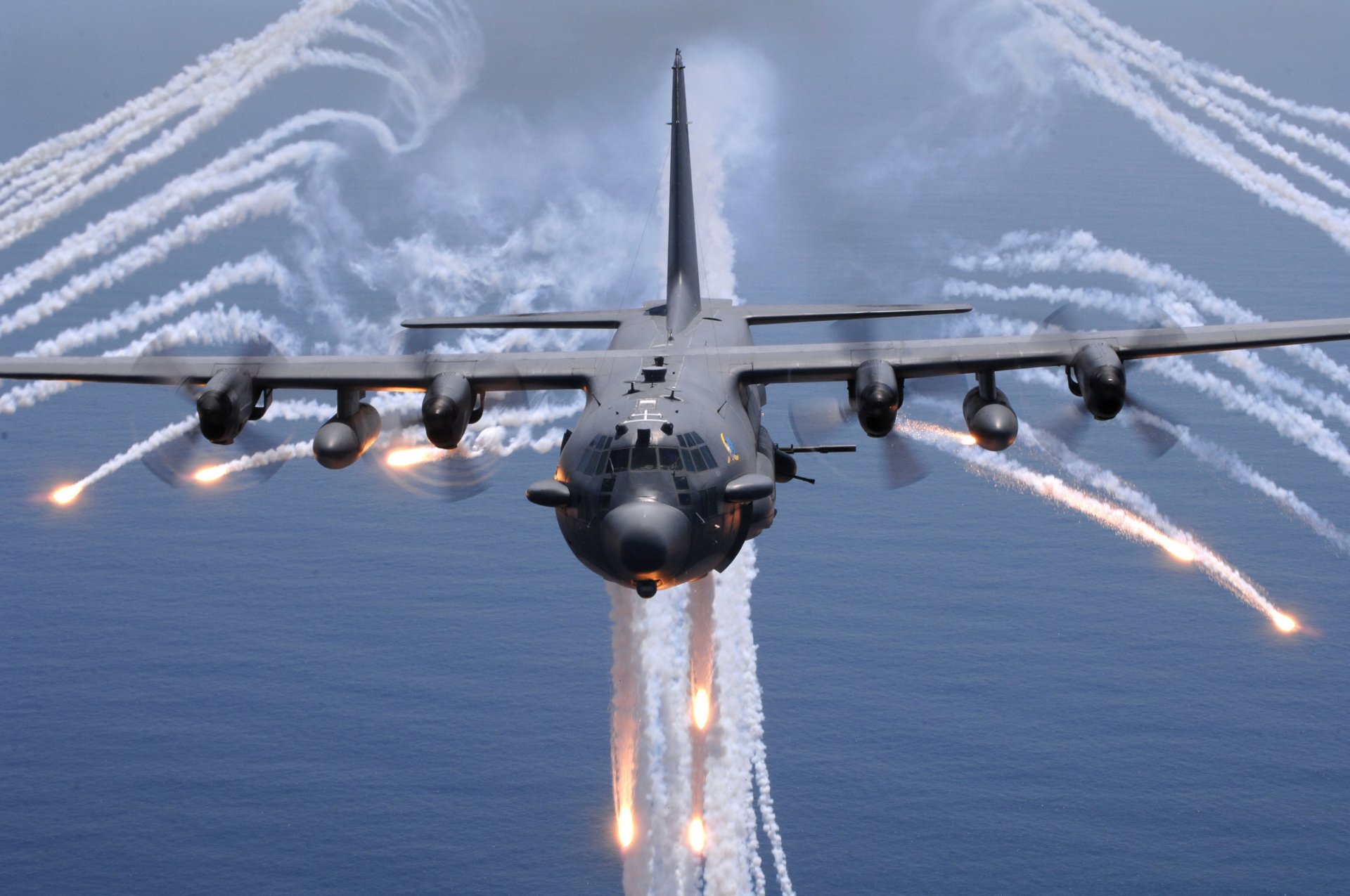 HD desktop wallpaper featuring a Lockheed AC-130 military aircraft releasing flares over the ocean under a cloudy sky.