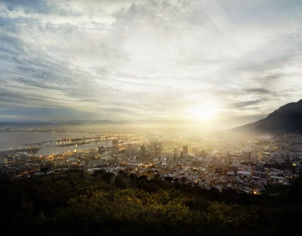 Panoramic night view of Cape Town, South Africa, with skyscrapers and city lights glowing through morning fog, framed by mountains and a cloudy sky.