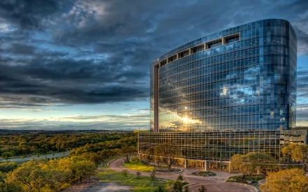 A striking HD desktop wallpaper featuring a modern, curved glass building against a dramatic sky, surrounded by lush greenery and a serene landscape.