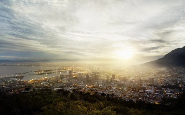 Panoramic night view of Cape Town, South Africa, with skyscrapers and city lights glowing through morning fog, framed by mountains and a cloudy sky.