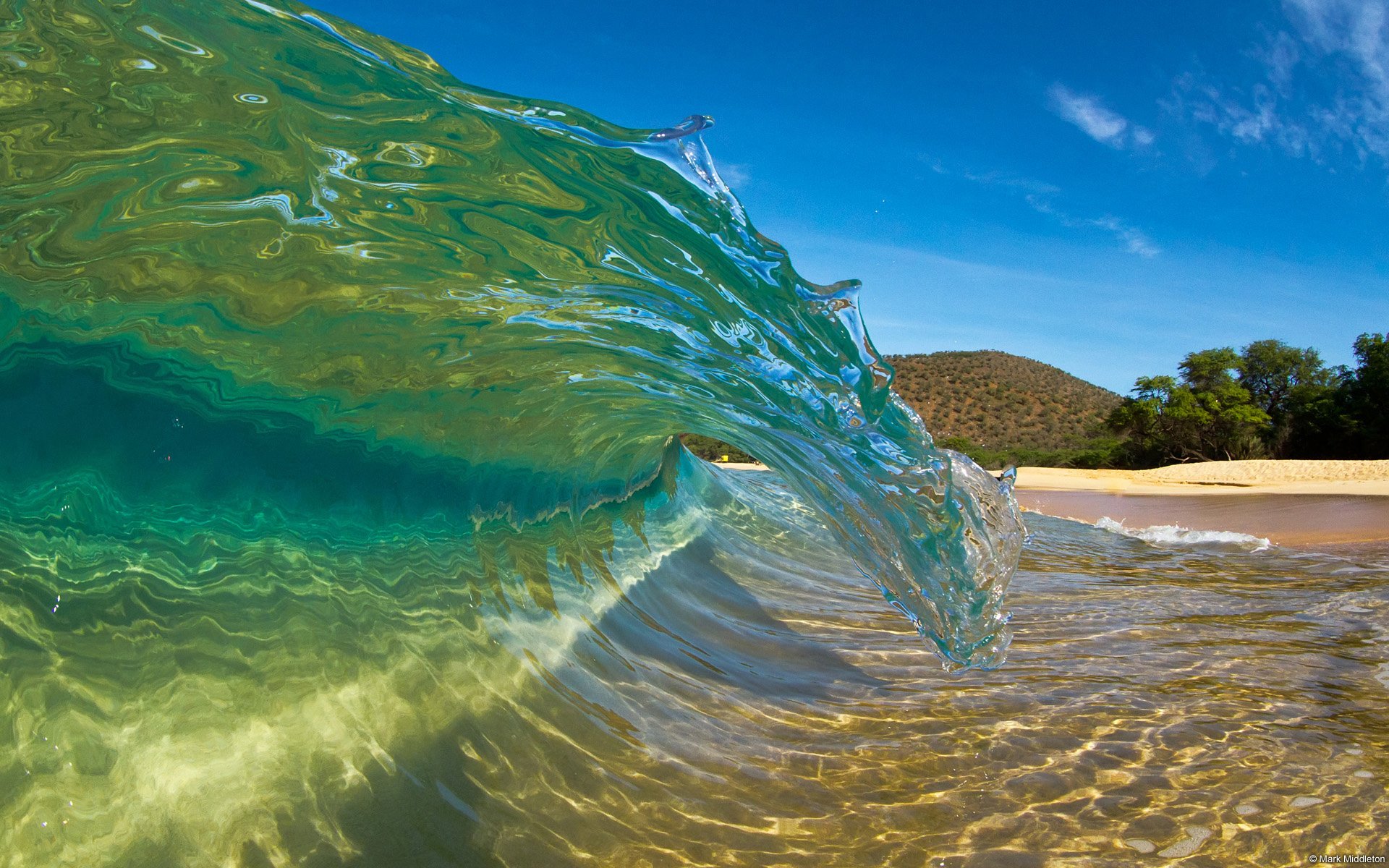 HD desktop wallpaper of a vibrant ocean wave curling near a sandy beach under a clear blue sky, capturing the dynamic beauty of nature.