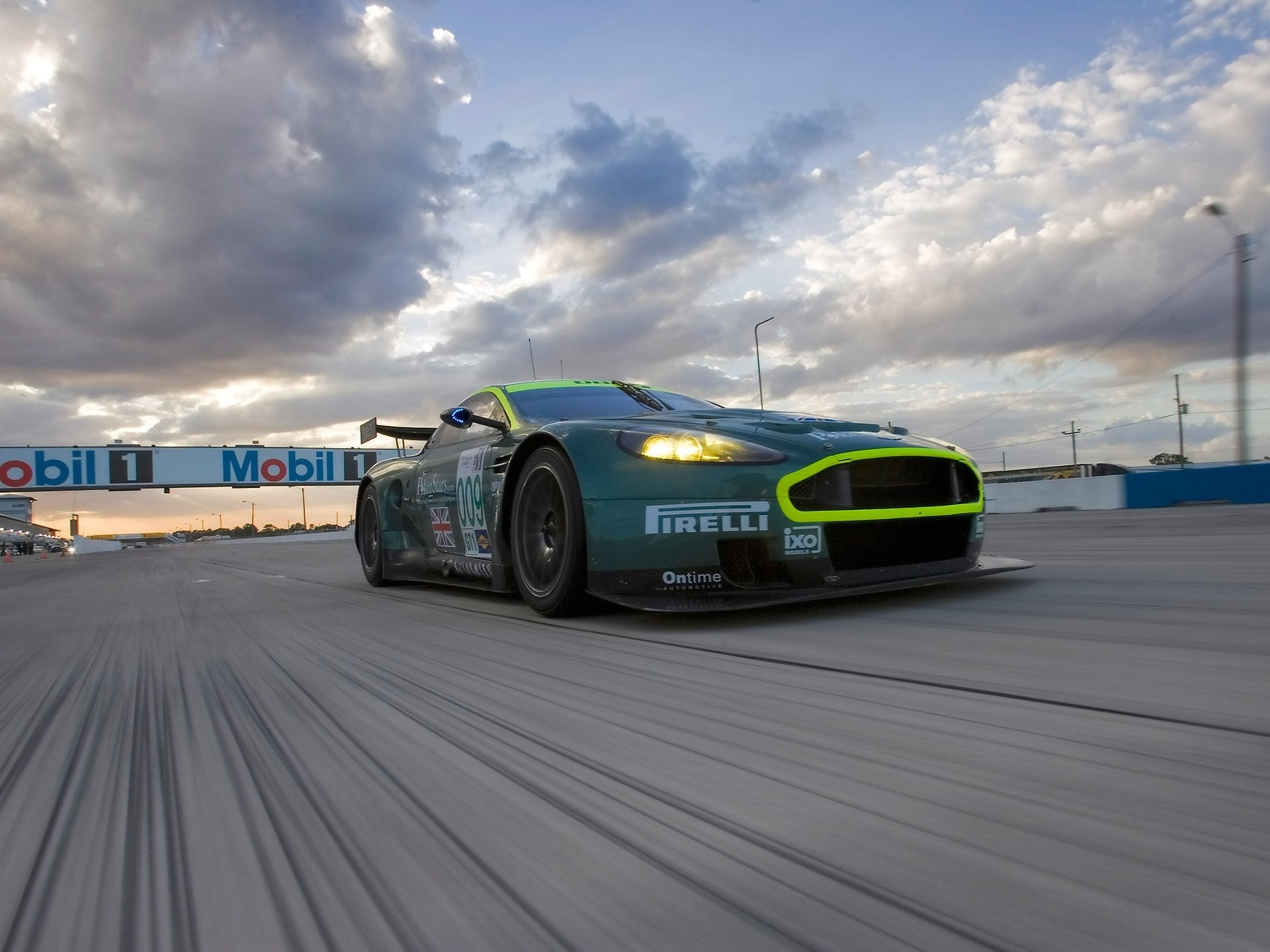 HD PC desktop wallpaper background: low-angle view of an Aston Martin DBR9 racing car speeding on a track beneath dramatic, cloud-filled sky.