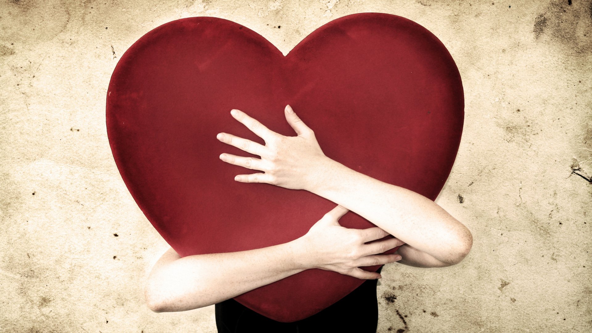 A hand embraces a large red heart against a textured background, capturing the essence of love in an artistic and intimate way. HD desktop wallpaper for a heartfelt touch.