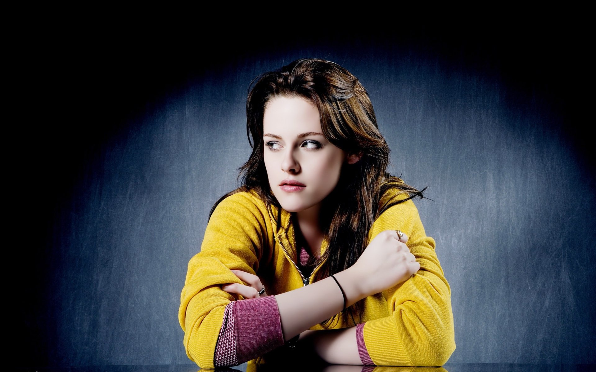 A striking HD wallpaper featuring Kristen Stewart, showcasing her thoughtful expression while wearing a bright yellow jacket against a moody background.