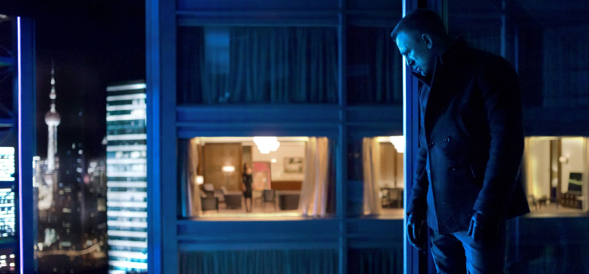 HD desktop wallpaper featuring Daniel Craig as James Bond in a moody scene from the movie Skyfall, with nighttime cityscape and illuminated windows in the background.