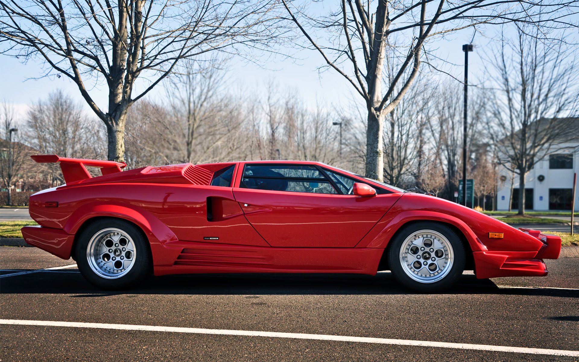 HD desktop wallpaper showcasing a vibrant red Lamborghini Countach parked outdoors on an asphalt surface with leafless trees and buildings in the background.