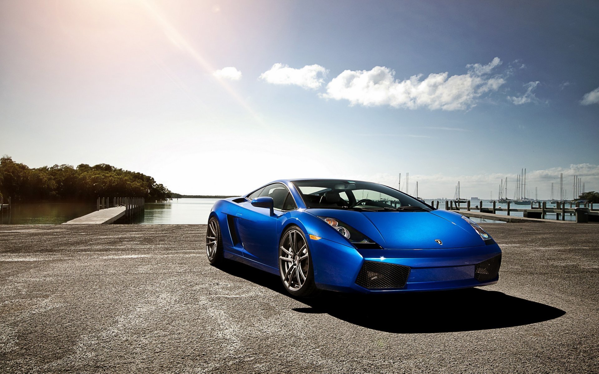 Blue Lamborghini Gallardo vehicle parked on a sunlit waterfront with marina and clear sky — 2K Quad HD PC desktop wallpaper and background.