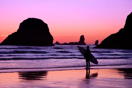 A serene sunset scene with a silhouetted surfer standing on the beach, surrounded by rocky formations and gentle waves, capturing the essence of surfing and sports in vibrant colors.