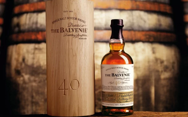 A beautifully displayed bottle of The Balvenie 40-Year-Old whisky, accompanied by its elegant wooden case, set against a backdrop of whiskey barrels, creating a rich food and drink atmosphere.