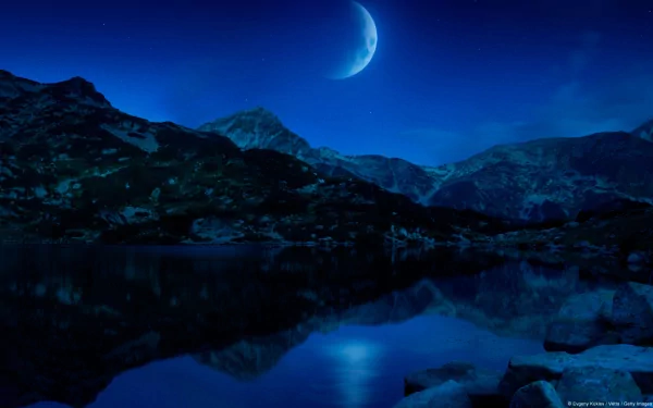 HD desktop wallpaper featuring a crescent moon glowing over a mountain landscape with a tranquil lake reflecting the night sky in a fantasy-inspired scenic photograph.