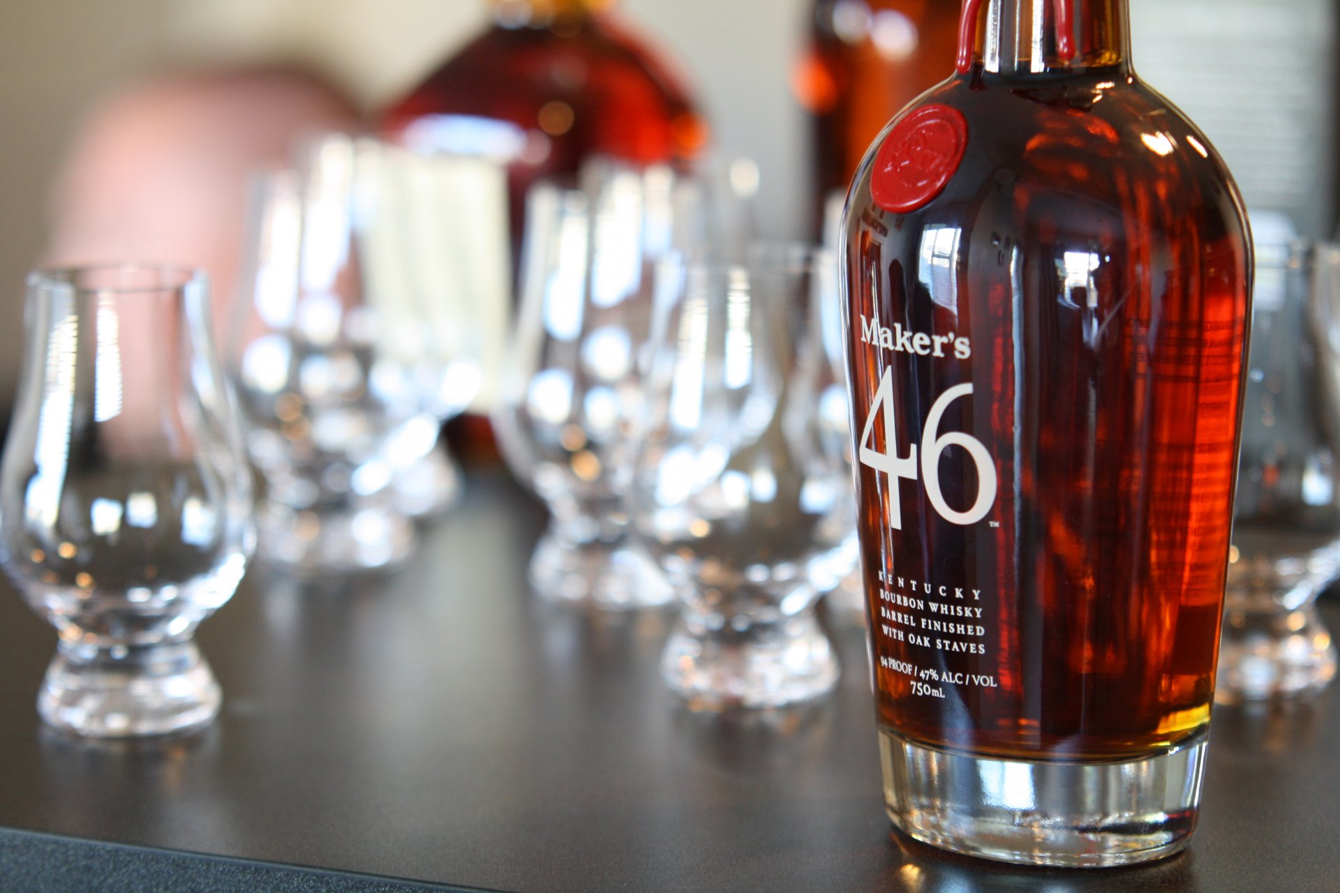 4K Ultra HD desktop wallpaper of a Maker's 46 whisky bottle in focus with tasting glasses and blurred bottles in the background, warm amber tones on a dark surface.