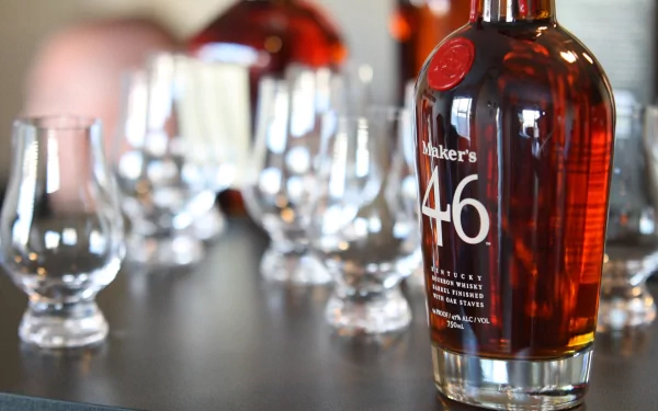 4K Ultra HD desktop wallpaper of a Maker's 46 whisky bottle in focus with tasting glasses and blurred bottles in the background, warm amber tones on a dark surface.