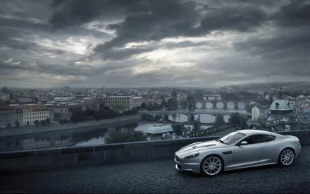 A stunning Aston Martin DBS parked on a scenic overlook, with a dramatic cloudy sky and a cityscape in the background, creating an elegant and captivating desktop wallpaper.