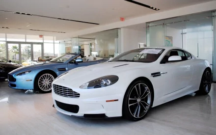 HD desktop wallpaper featuring two Aston Martin vehicles displayed in a modern showroom with polished floors and large glass windows.