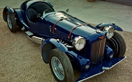 HD PC desktop wallpaper featuring a classic dark blue Aston Martin vintage race car with leather straps and prominent front grille, parked on a gravel surface.