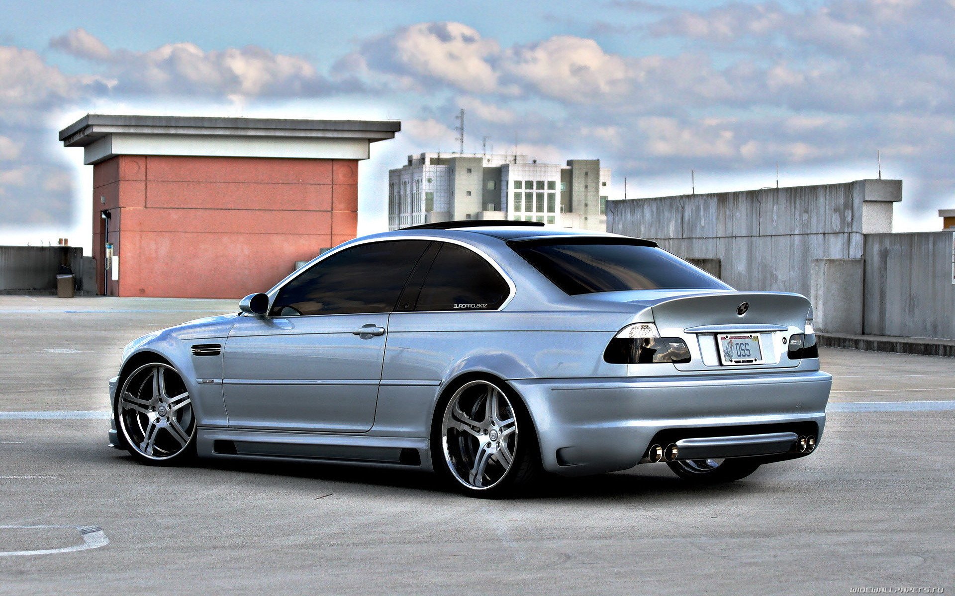 HD PC desktop wallpaper featuring a sleek silver BMW vehicle parked on a rooftop with a cityscape and cloudy sky in the background.