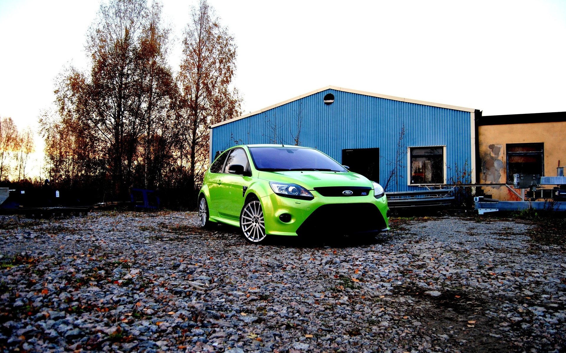 A vibrant green Ford Focus RS500 parked on a gravel surface near blue and beige industrial buildings, captured in an HD PC desktop wallpaper background.