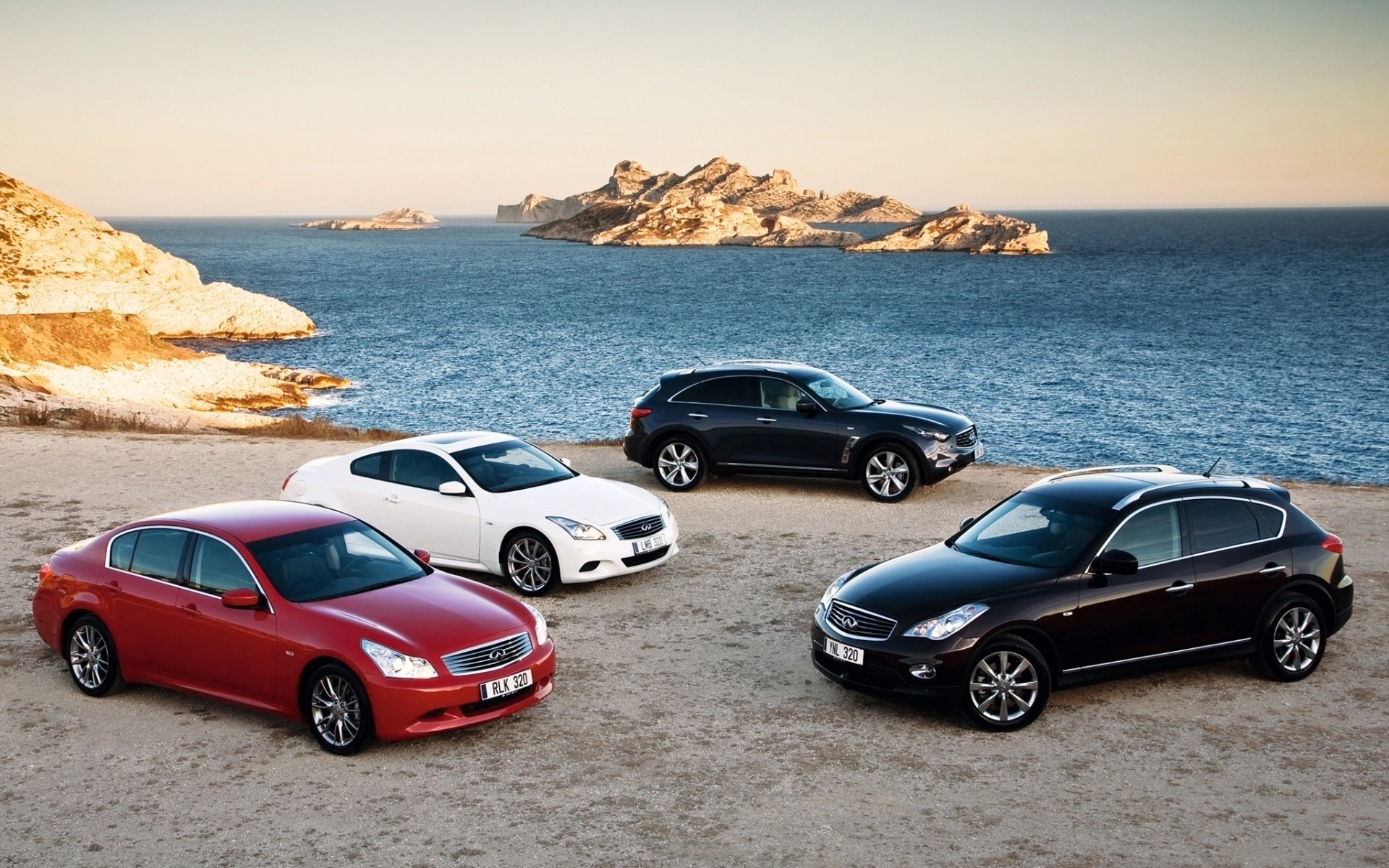 HD PC desktop wallpaper showing four Infiniti vehicles—a red sedan, white coupe, black crossover and dark hatchback—parked on a sandy coastal shore with rocky islands and blue sea.