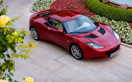 HD PC desktop wallpaper and background showing a glossy red Lotus sports car parked on a stone driveway beside manicured flowerbeds, seen from above.