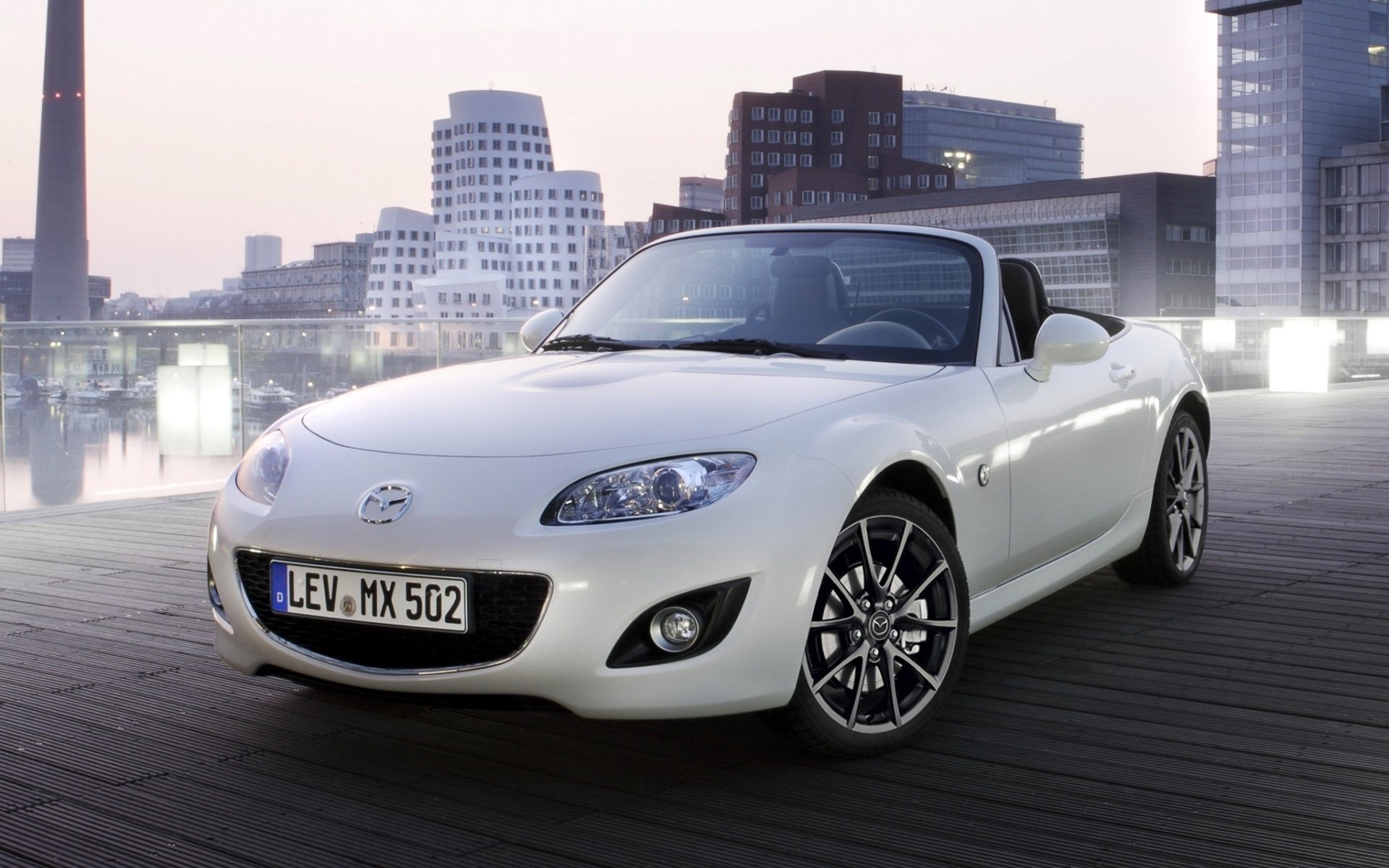 White Mazda convertible on a waterfront boardwalk with city skyline at dusk — HD PC desktop wallpaper and background.