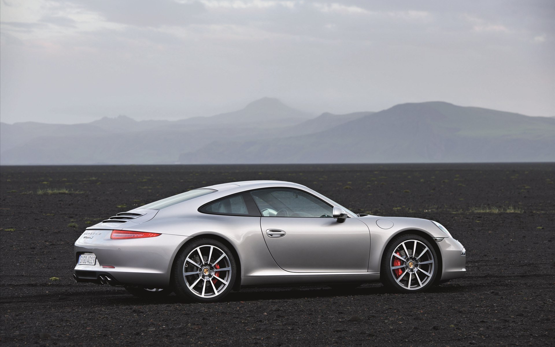 Silver Porsche sports coupe on a black volcanic plain with distant mountains — HD PC desktop wallpaper and background.