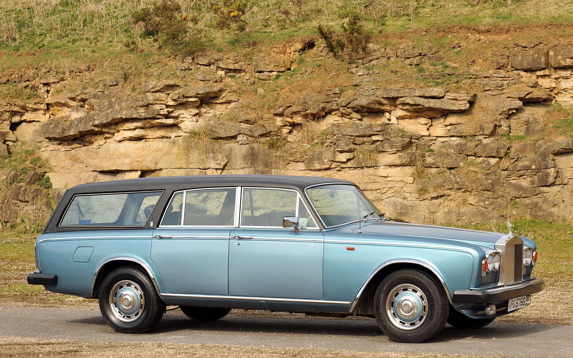 HD PC desktop wallpaper and background: light blue Rolls‑Royce estate car parked beside a rocky hillside, chrome grille and Spirit of Ecstasy hood ornament clearly visible.