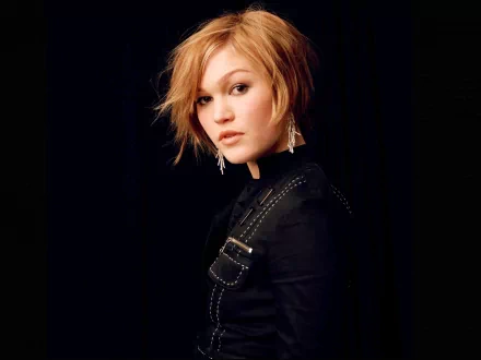 A high-definition desktop wallpaper featuring celebrity Julia Stiles with short blonde hair, wearing a black outfit and earrings, set against a dark background.