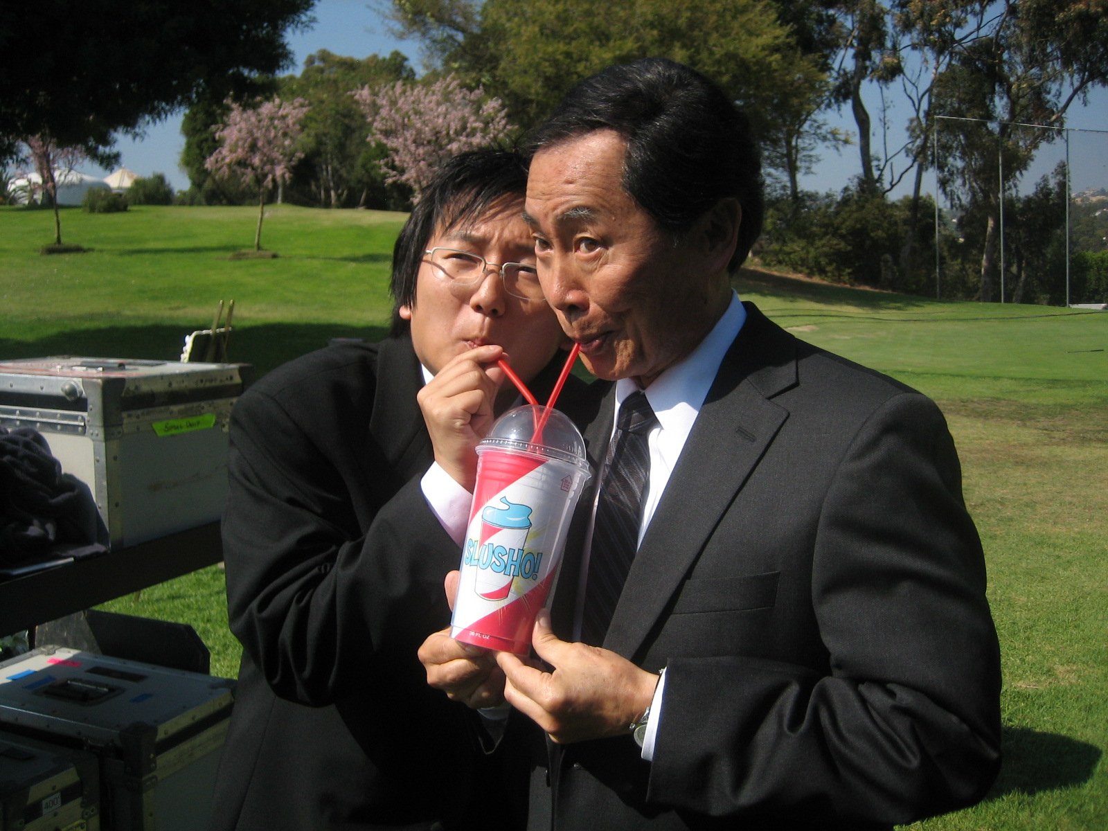 Two actors from the TV show Heroes share a fun moment, sipping from a large drink, while dressed in suits against a sunny outdoor backdrop. This serves as a vibrant desktop wallpaper.