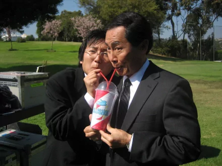 Two actors from the TV show Heroes share a fun moment, sipping from a large drink, while dressed in suits against a sunny outdoor backdrop. This serves as a vibrant desktop wallpaper.