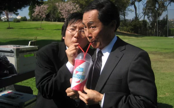 Two actors from the TV show Heroes share a fun moment, sipping from a large drink, while dressed in suits against a sunny outdoor backdrop. This serves as a vibrant desktop wallpaper.