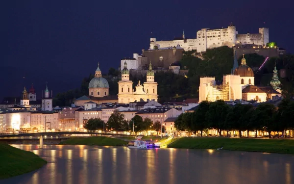 A stunning view of Hohensalzburg Castle illuminated at night, overlooking the Salzburg skyline with a serene river reflecting the city's historic architecture.