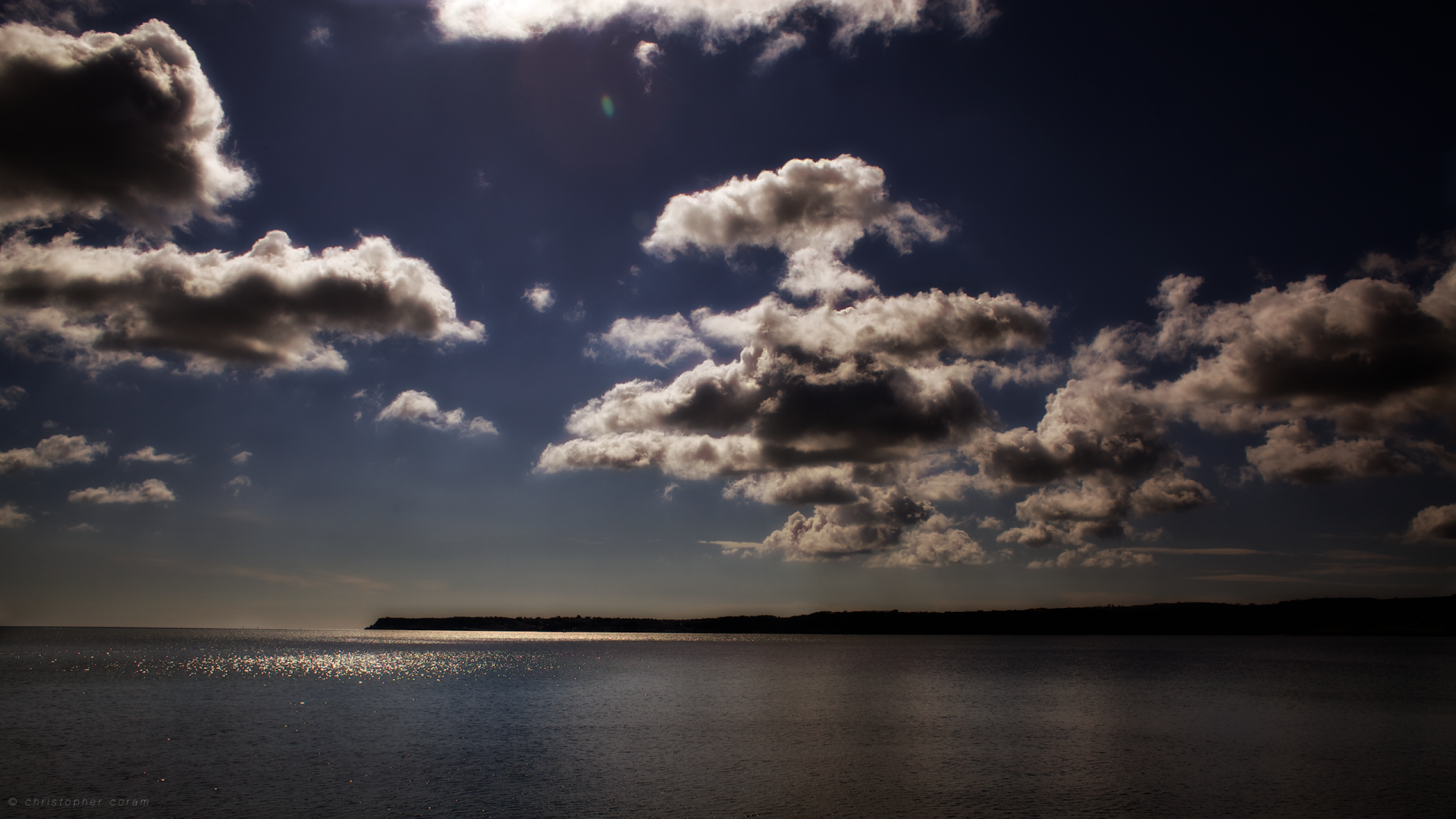 A serene view of a dark blue sky filled with fluffy clouds, reflecting over a calm sea, capturing the tranquil beauty of nature under the sun. Ideal HD wallpaper for desktop backgrounds.