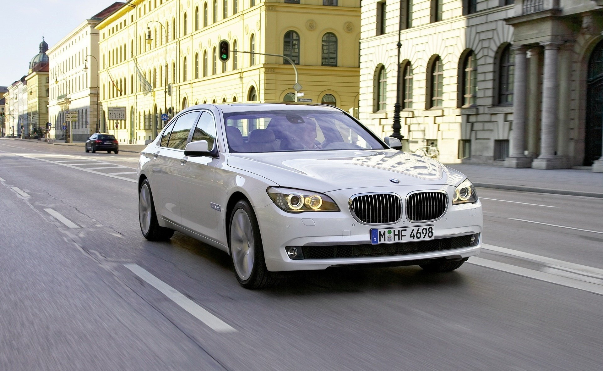 HD PC desktop wallpaper of a white BMW vehicle sedan driving down a sunlit European street, motion blur and classic architecture forming the background.