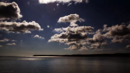A serene view of a dark blue sky filled with fluffy clouds, reflecting over a calm sea, capturing the tranquil beauty of nature under the sun. Ideal HD wallpaper for desktop backgrounds.