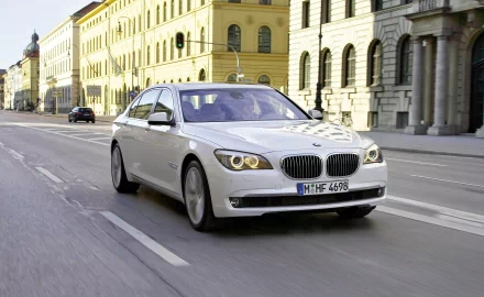 HD PC desktop wallpaper of a white BMW vehicle sedan driving down a sunlit European street, motion blur and classic architecture forming the background.